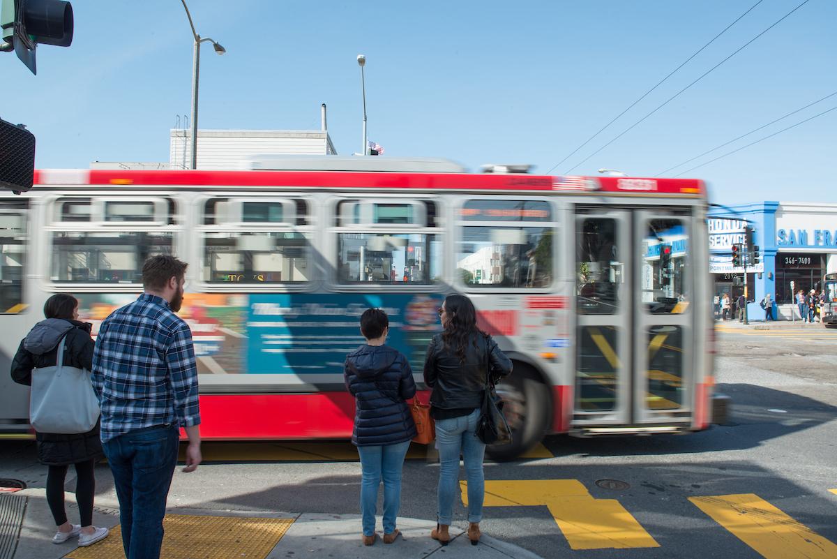 People wait to cross the street as a bus passes in front of them in an intersection. People wait to cross the street as a bus passes in front of them in an intersection.